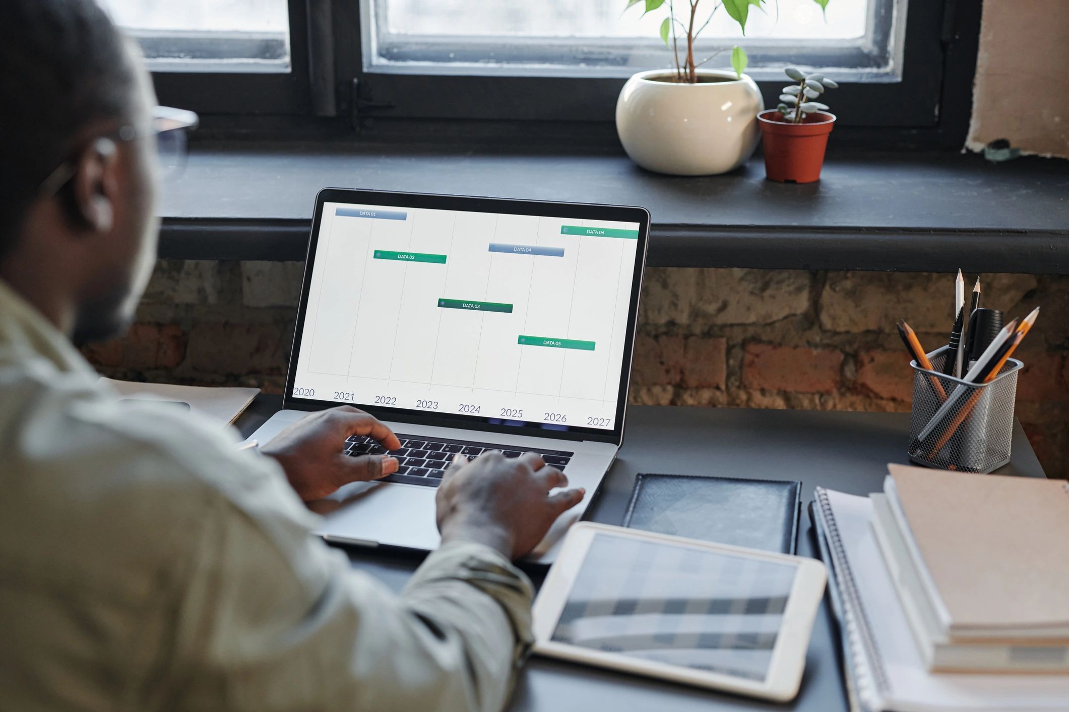 Horizontal medium shot of unrecognizable young Black man sitting at desk in office working with stock trading data using laptop